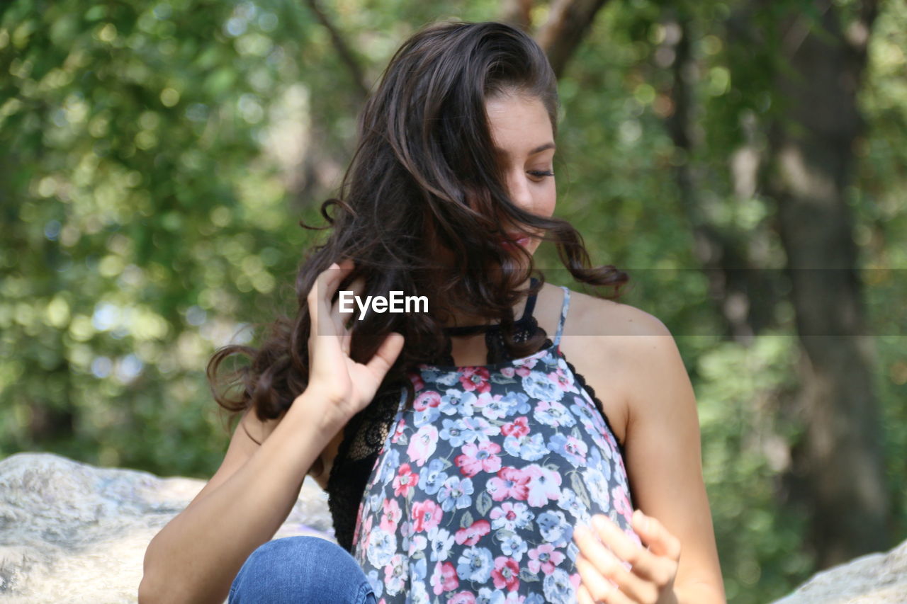 Beautiful young woman with hand in hair sitting on rocks against trees