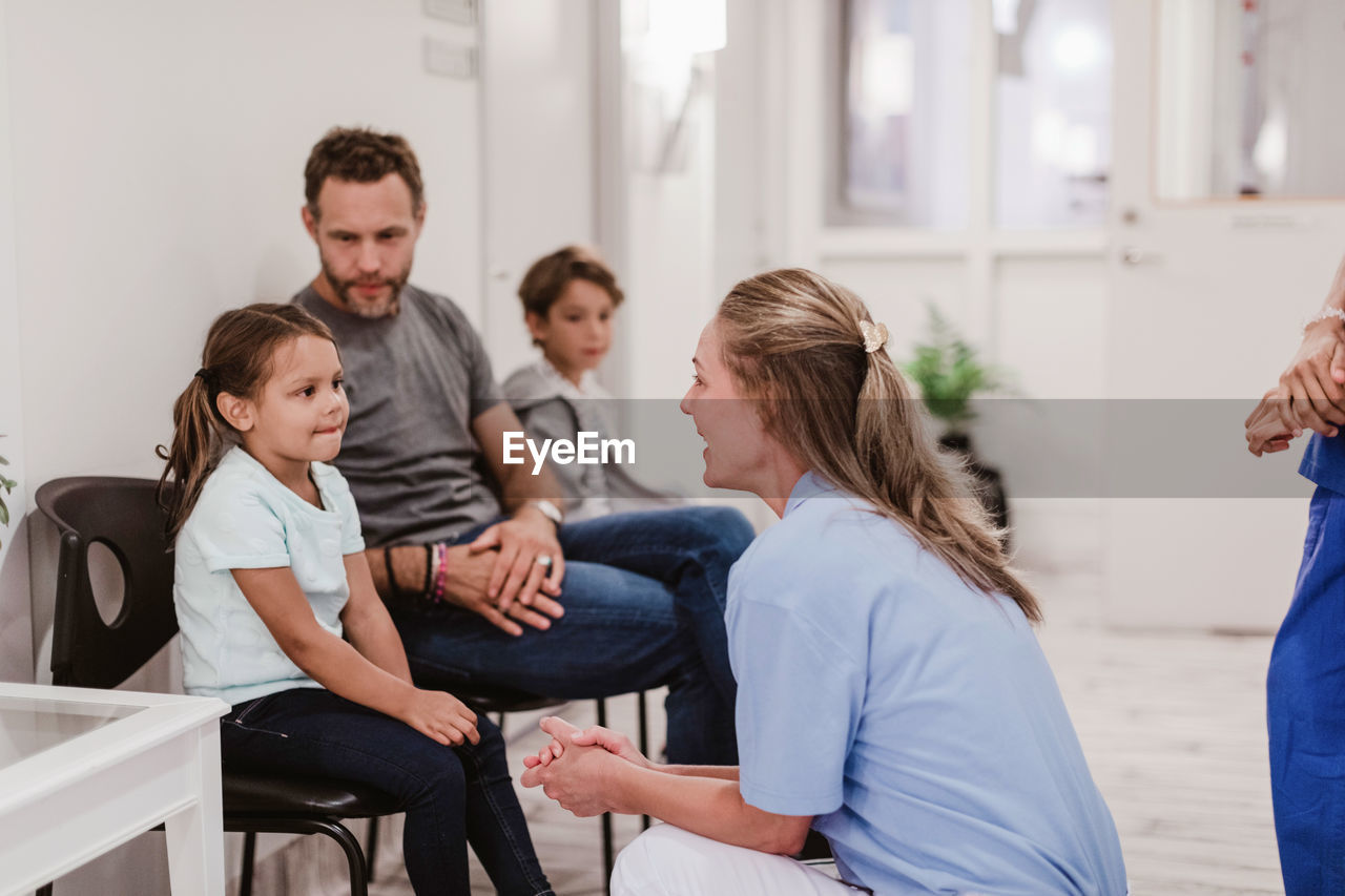 Female pediatrician talking with girl sitting by family in hospital corridor