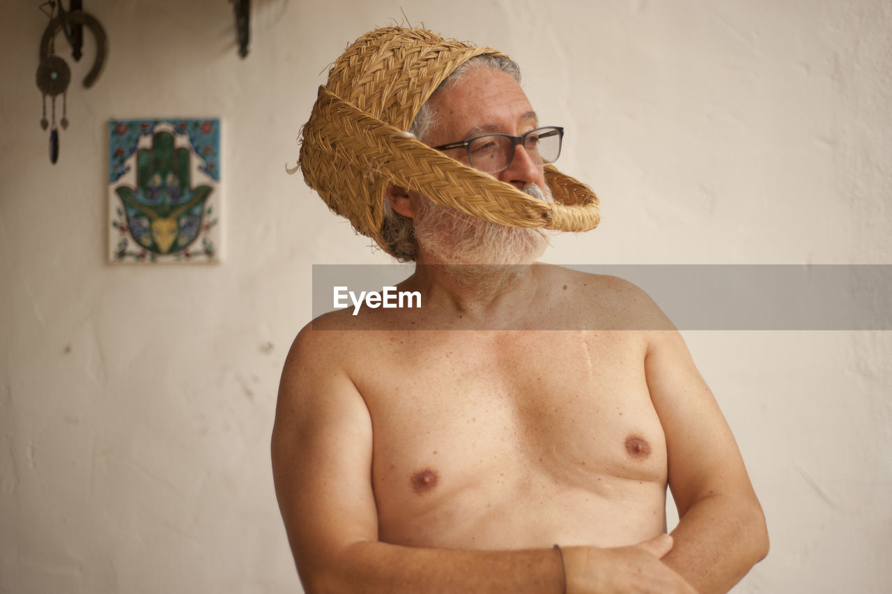 Shirtless man wearing basket while standing against wall