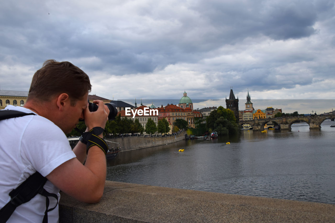 Photographer photographing bridge while leaning on railing against cloudy sky