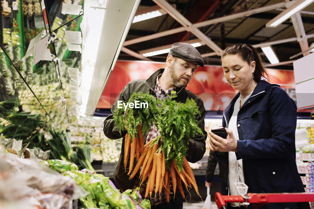 Couple using smart phone while buying carrot at supermarket