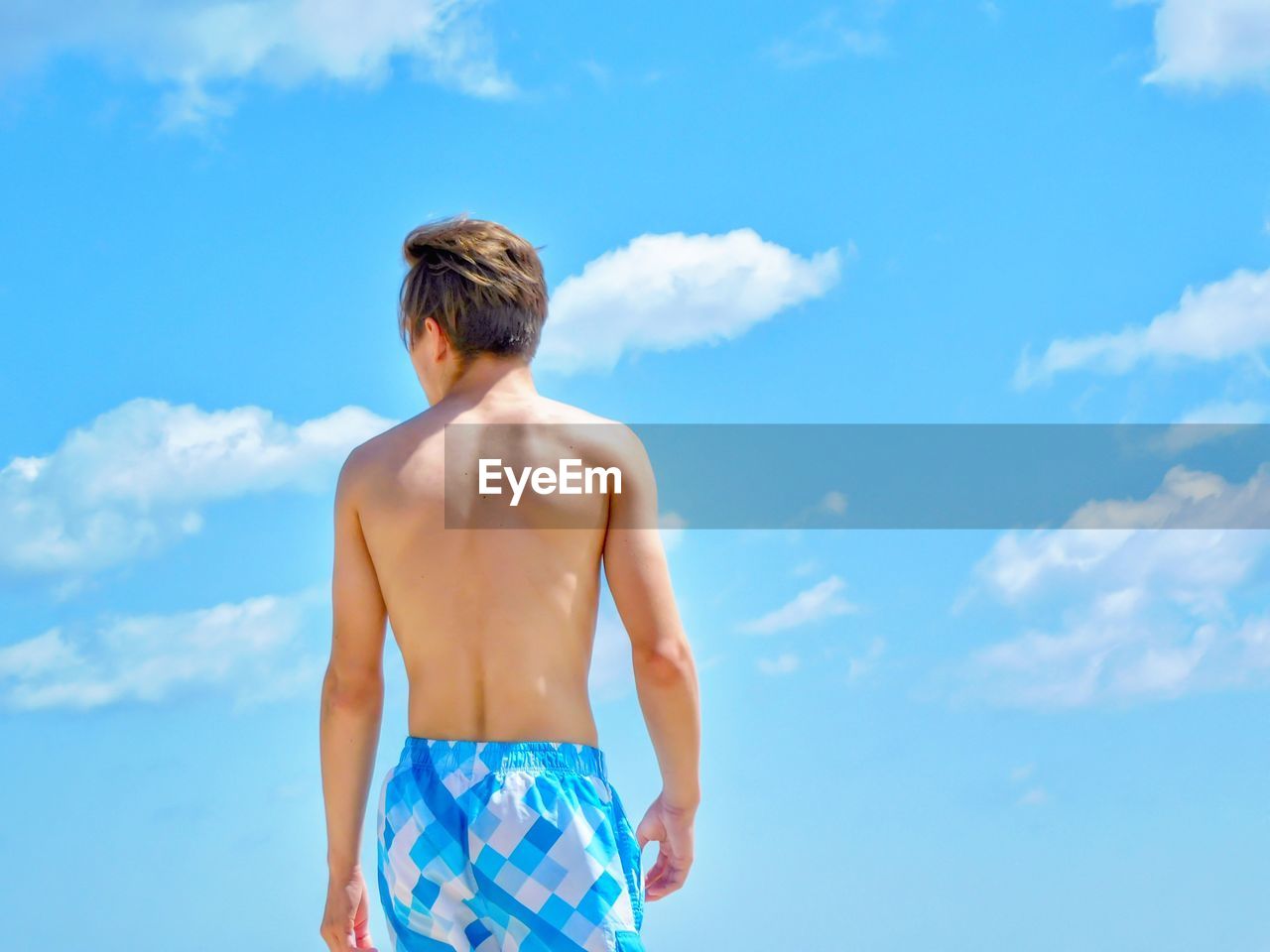 Rear view of shirtless teenage boy standing against blue sky