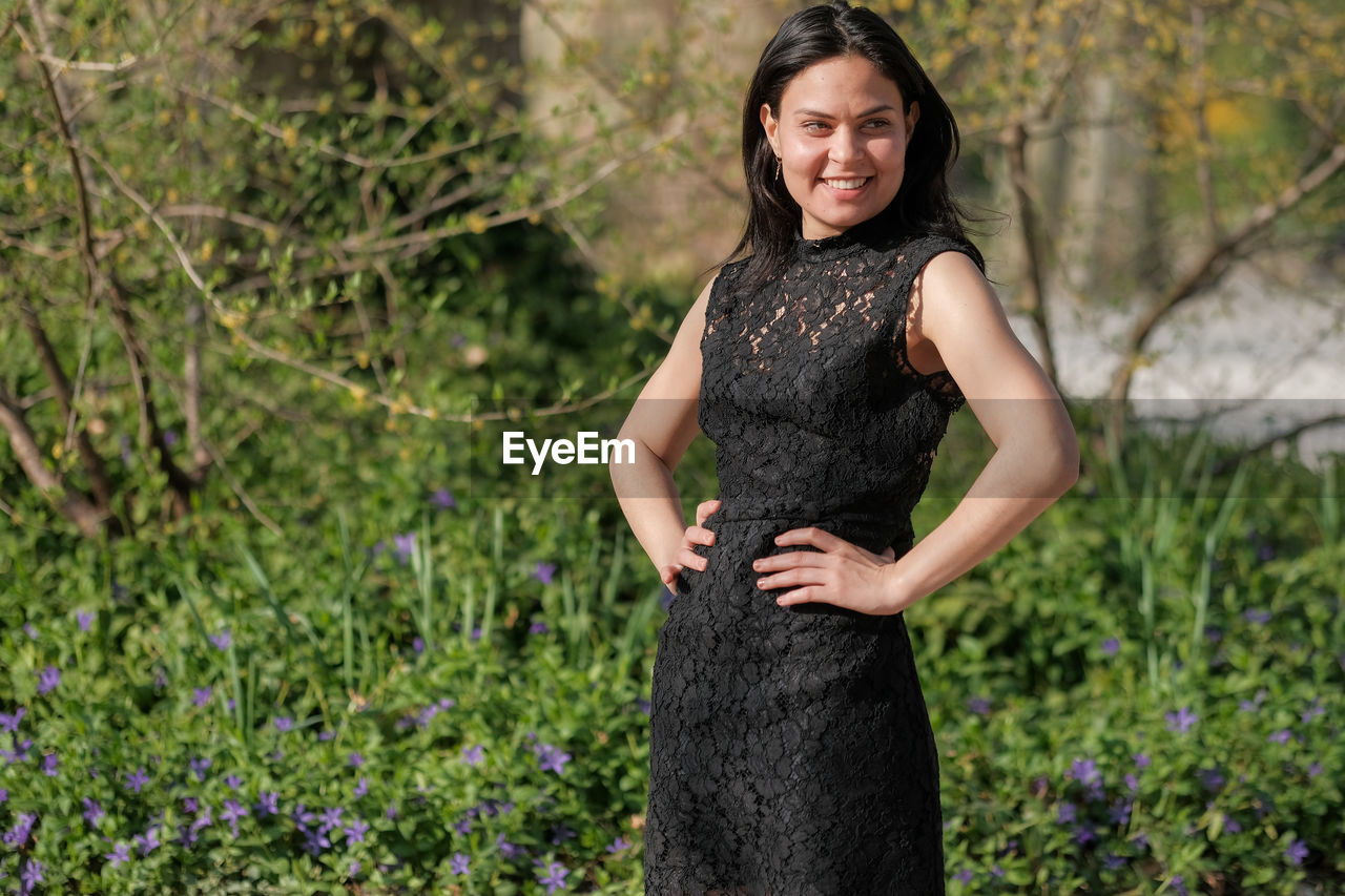 one person, plant, women, adult, portrait, smiling, nature, dress, looking at camera, three quarter length, standing, happiness, flower, hairstyle, clothing, flowering plant, young adult, emotion, fashion, woodland, female, land, outdoors, long hair, lifestyles, green, spring, beauty in nature, front view, black hair, focus on foreground, photo shoot, grass, day, tree, brown hair, cheerful, growth, hand, person, portrait photography