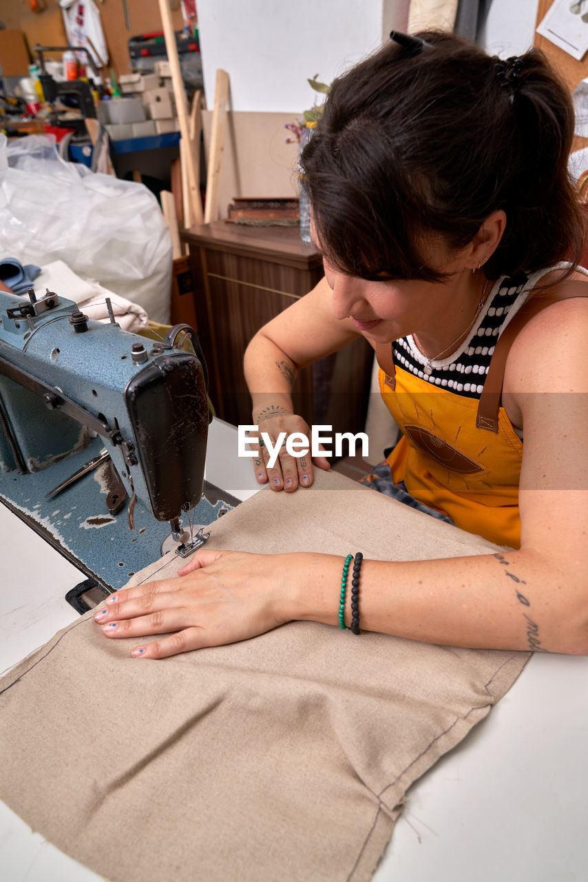 From above of positive tattooed adult female artisan in apron with dark hair sewing fabric with machine while working in traditional workshop