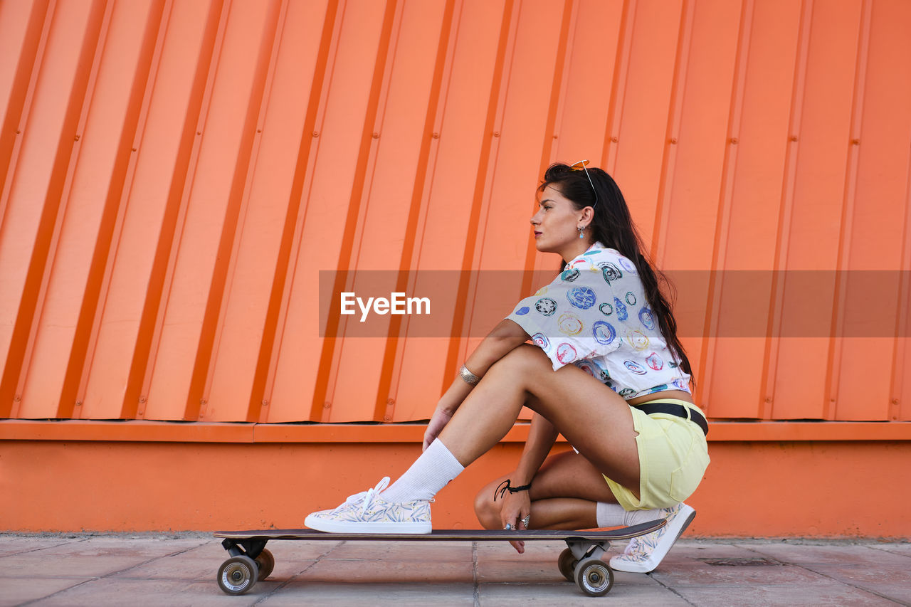 Portrait of beautiful girl crouching beside longboard in front of corrugated iron wall