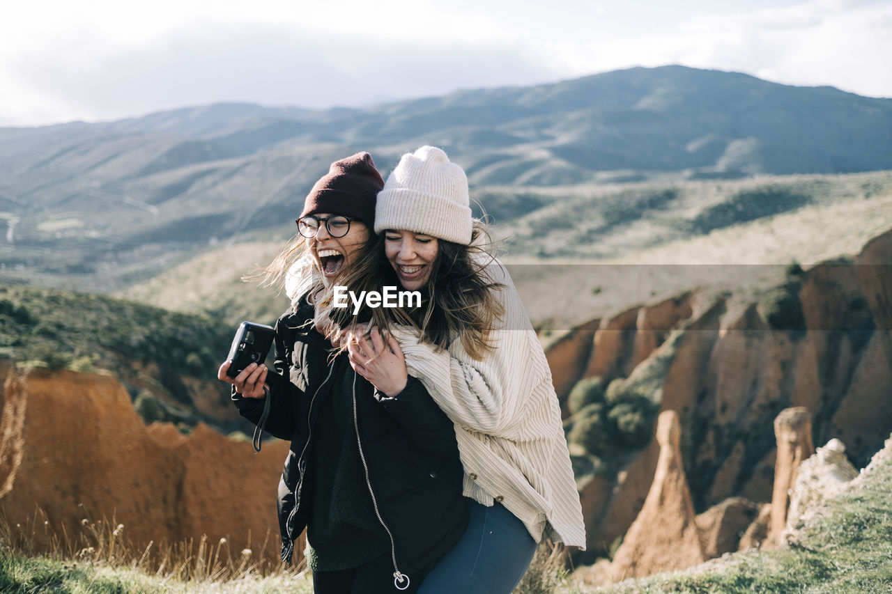 Cheerful best female friends with photo camera having fun against mount and gorge on sunny day in back lit