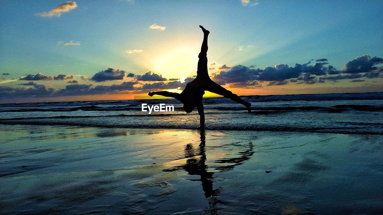 Man doing handstand at beach against sky during sunset