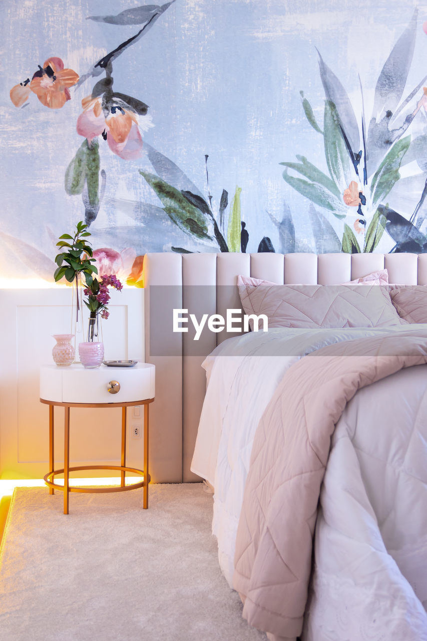 Interior of bedroom with floral ornament on wall with comfortable bed under blanket placed near round bedside table with vase of fresh plants