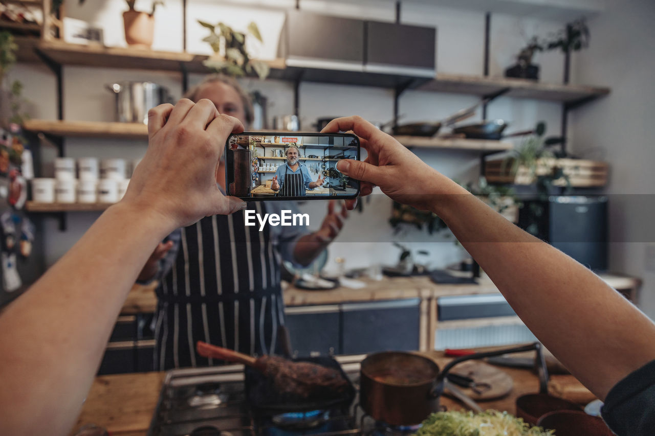 Young woman filming mature chef through mobile phone while standing in kitchen