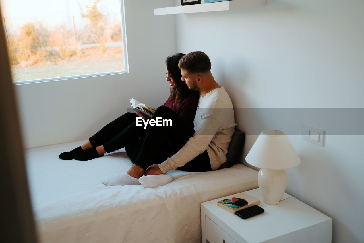 Side view of tender couple sitting on comfortable bed and reading interesting book together