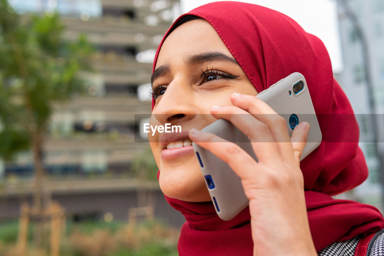 Low angle of positive ethnic female in hijab standing on street speaking on smartphone enjoying weekend