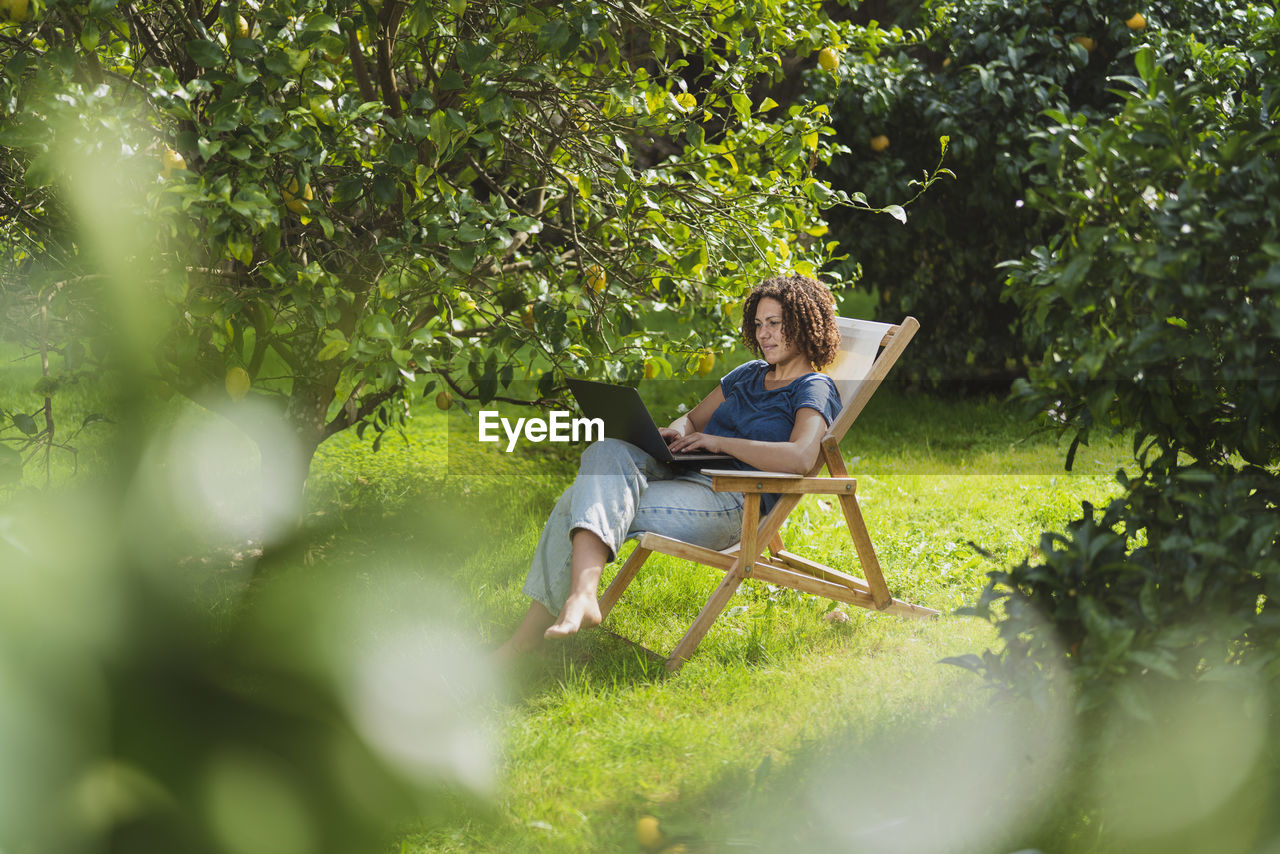 Mid adult woman using laptop while sitting on chair amidst trees in garden