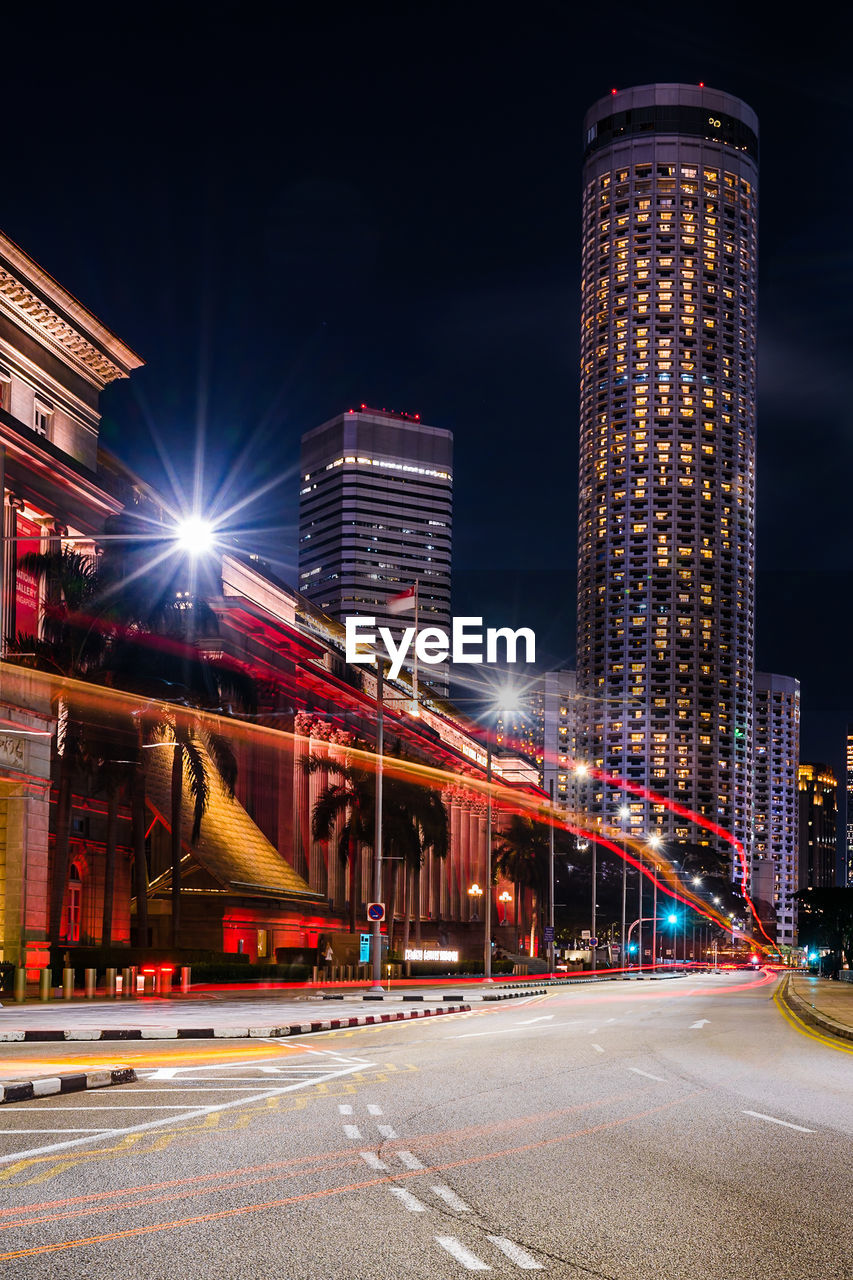 Light trails on road by illuminated buildings against sky at night