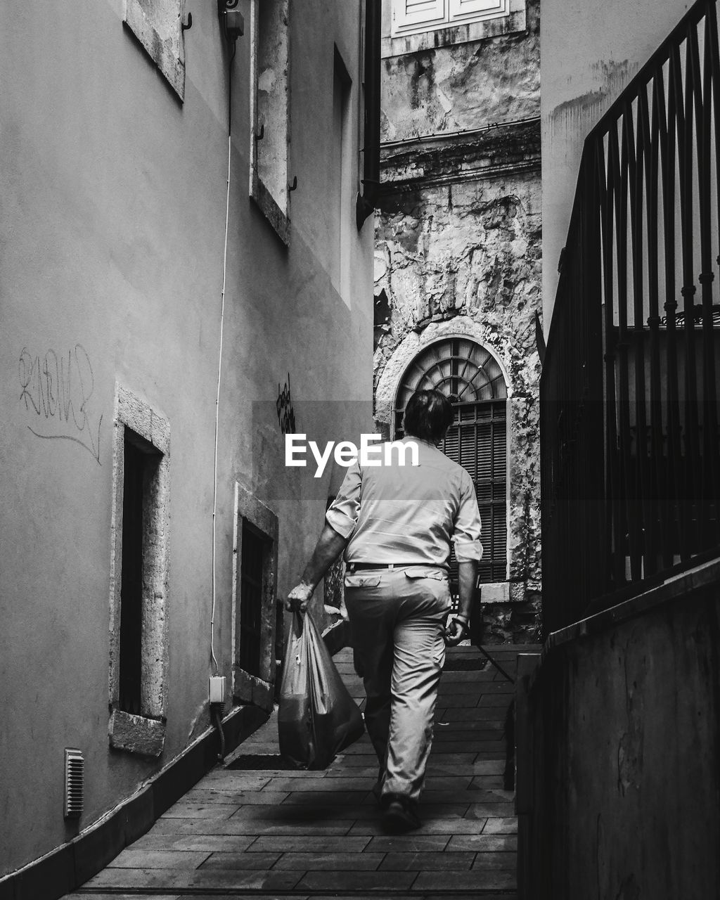 REAR VIEW OF MAN WALKING IN ALLEY AMIDST BUILDINGS