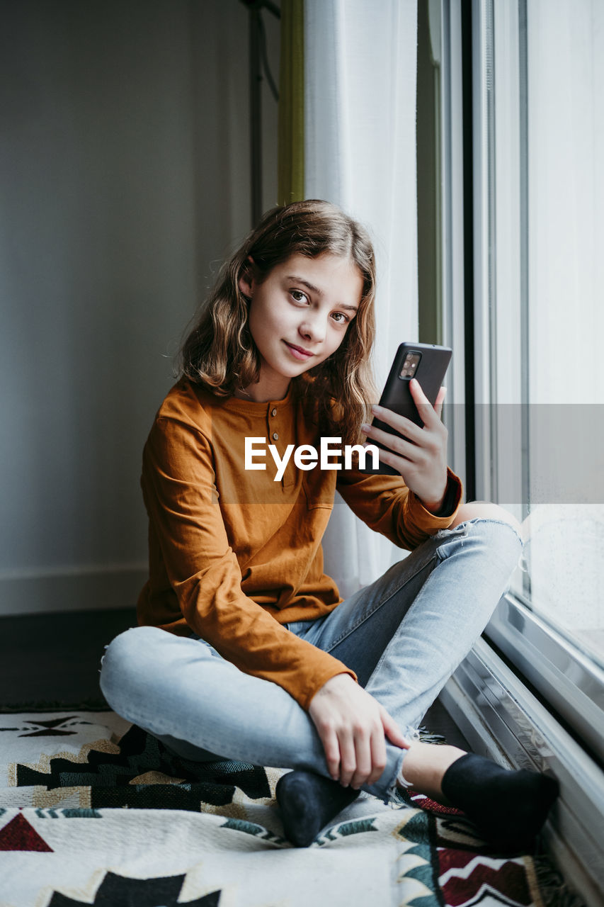 Girl with mobile phone sitting cross legged by window at home