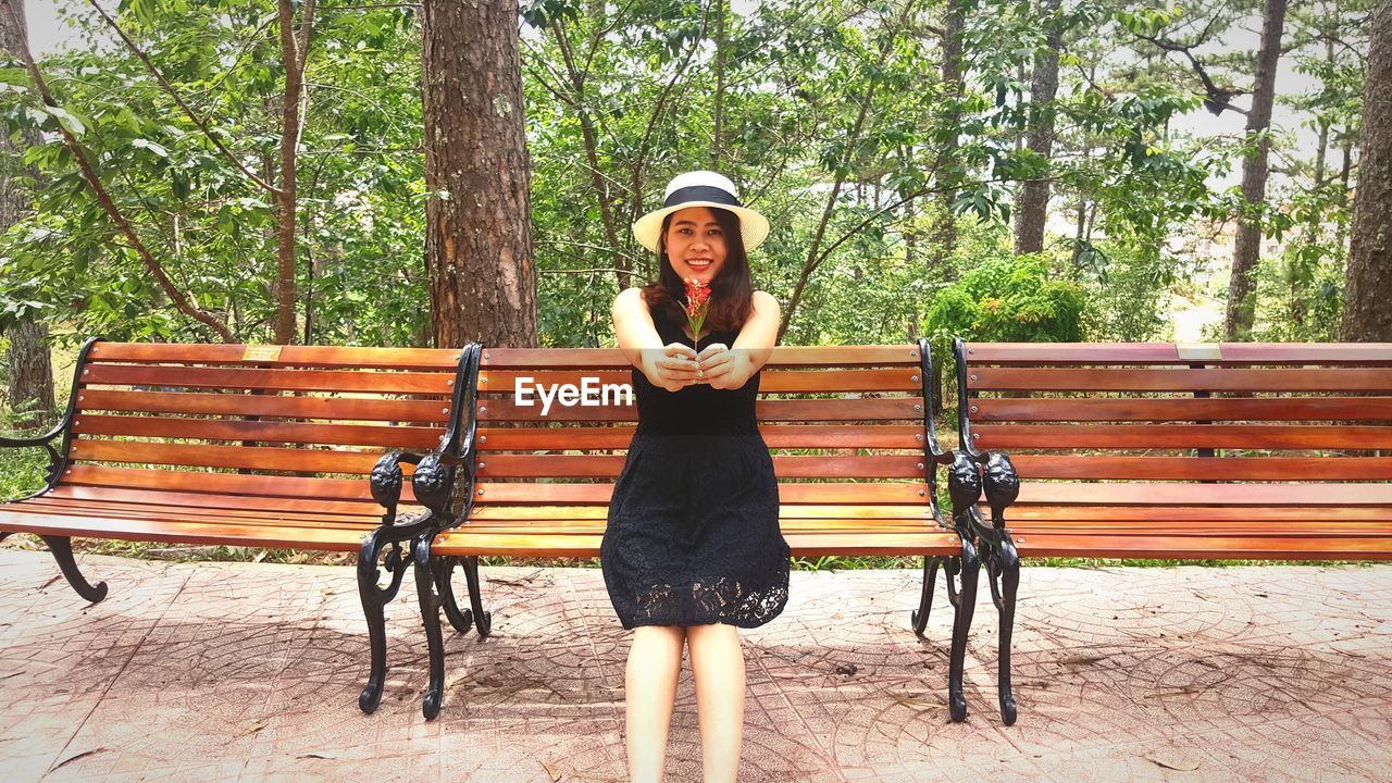 Portrait of young woman giving flowers while sitting on bench at park