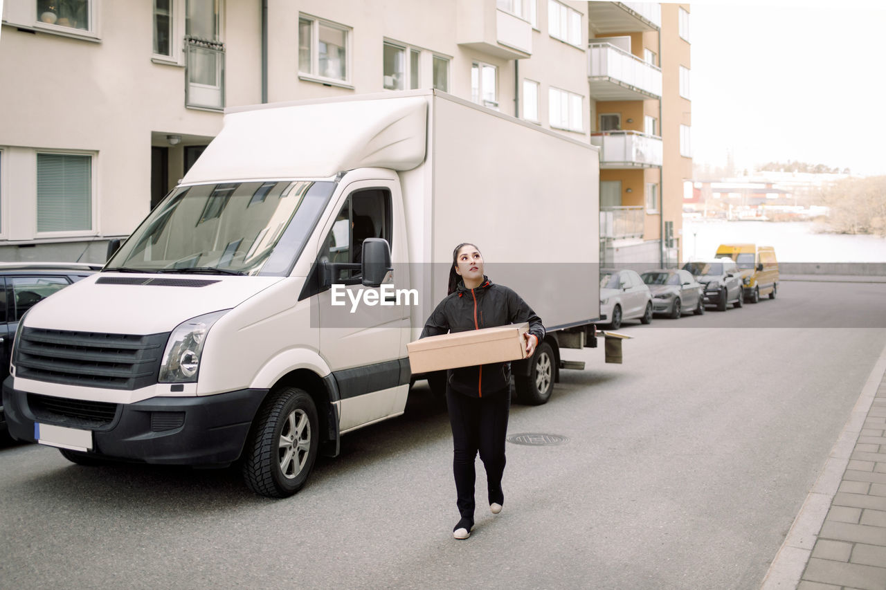 Young delivery woman carrying box while walking on street in city