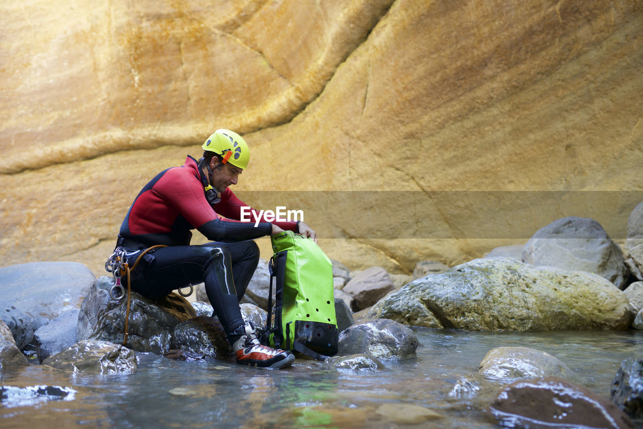 Side view of male hiker with backpack sitting on rocks in river