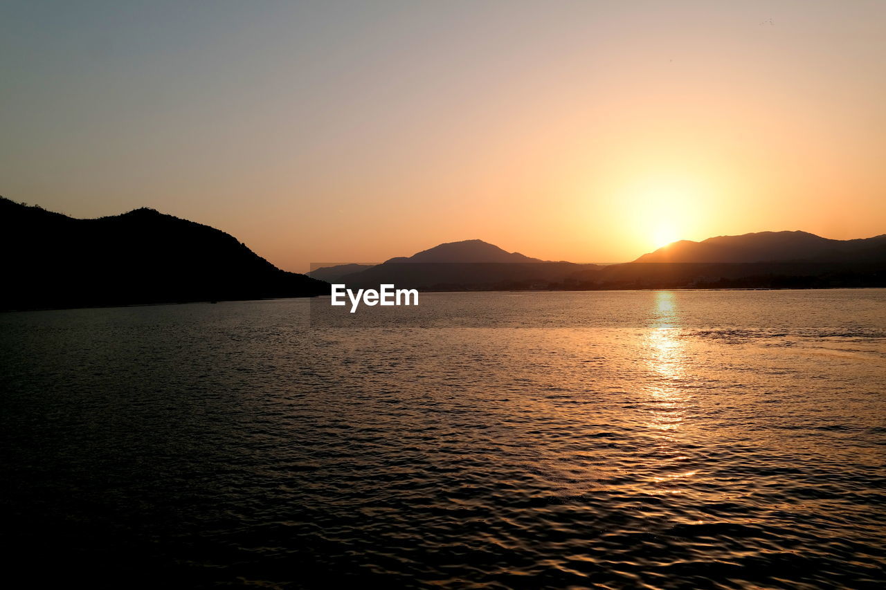 SCENIC VIEW OF SEA BY SILHOUETTE MOUNTAINS AGAINST SKY DURING SUNSET