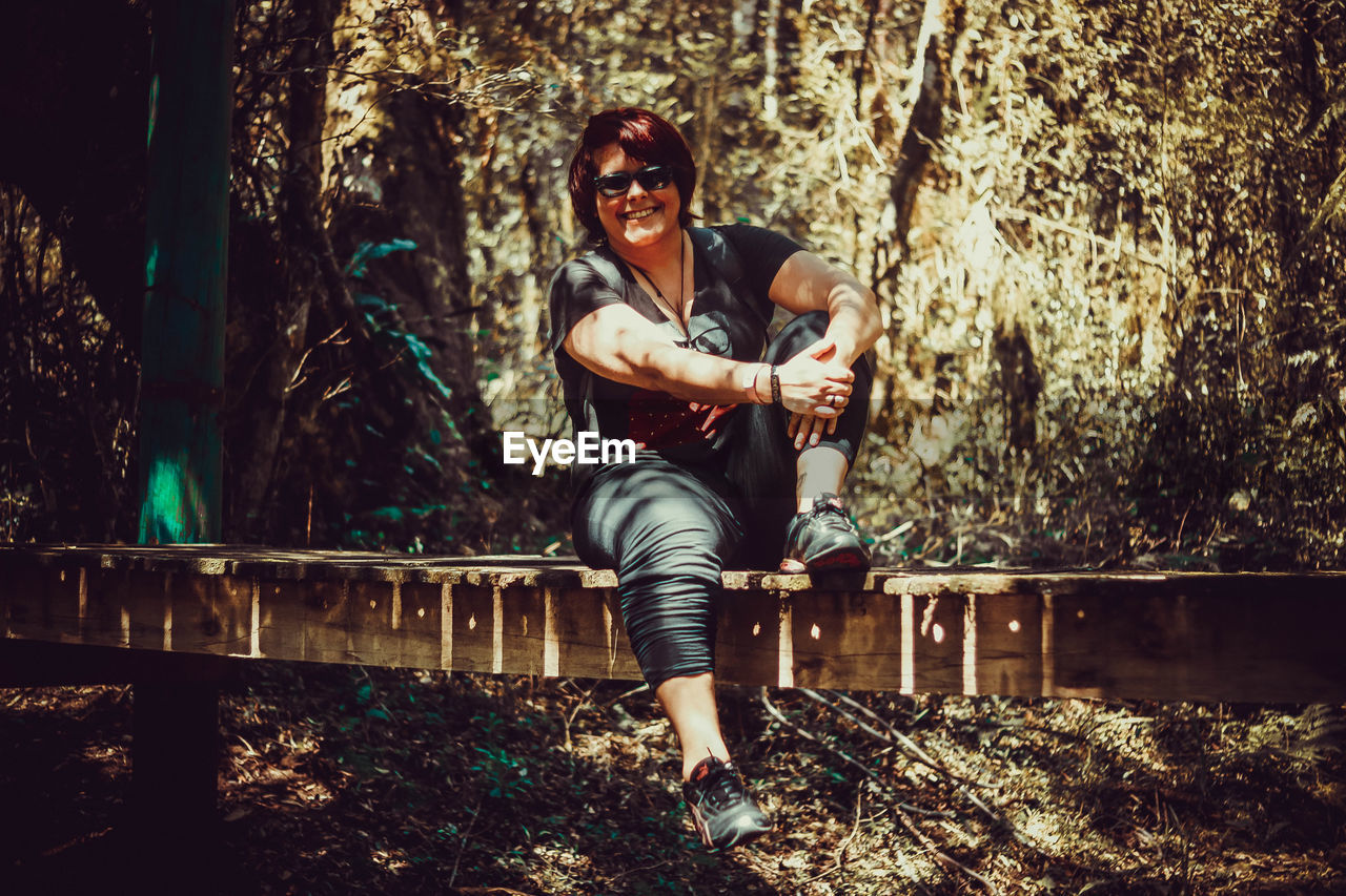 Portrait of smiling woman wearing sunglasses sitting on footbridge in forest