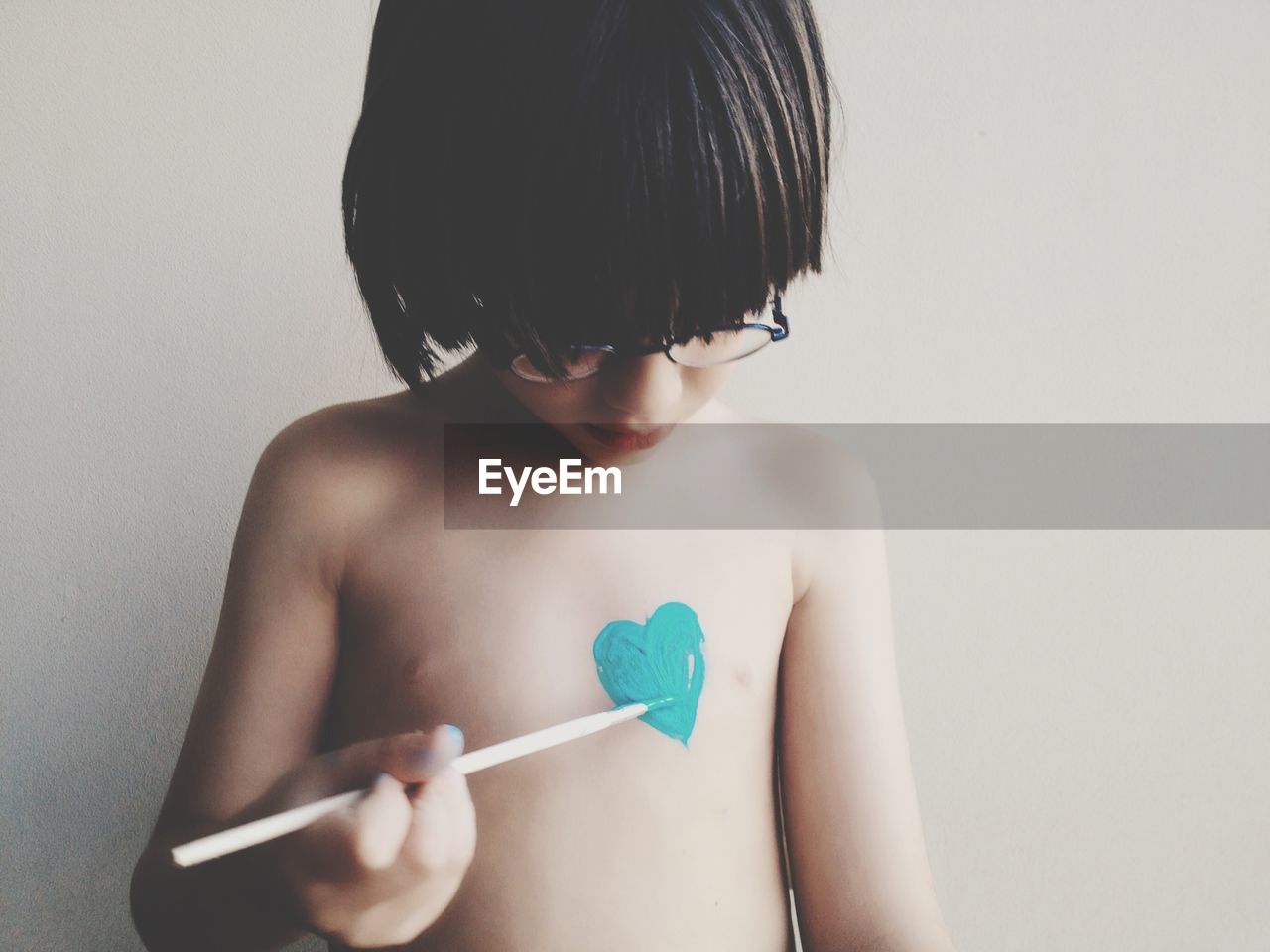 Shirtless boy painting heart shape on chest at home