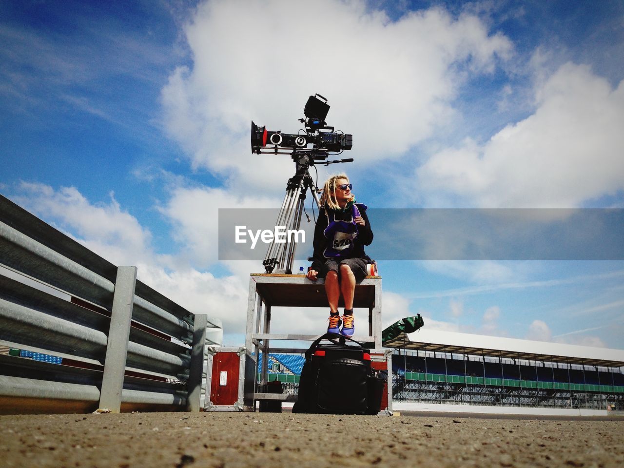 Full length of female operator sitting by camera in stadium against sky