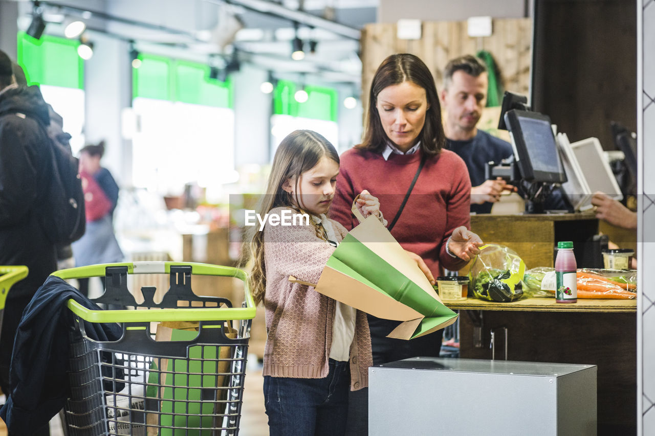 Daughter holding paper bag while standing by mother at checkout counter in supermarket
