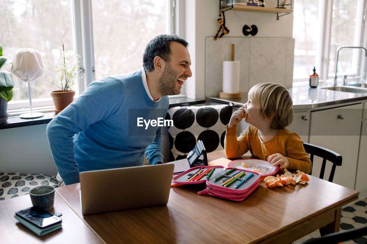 Playful father looking at daughter while using laptop at home