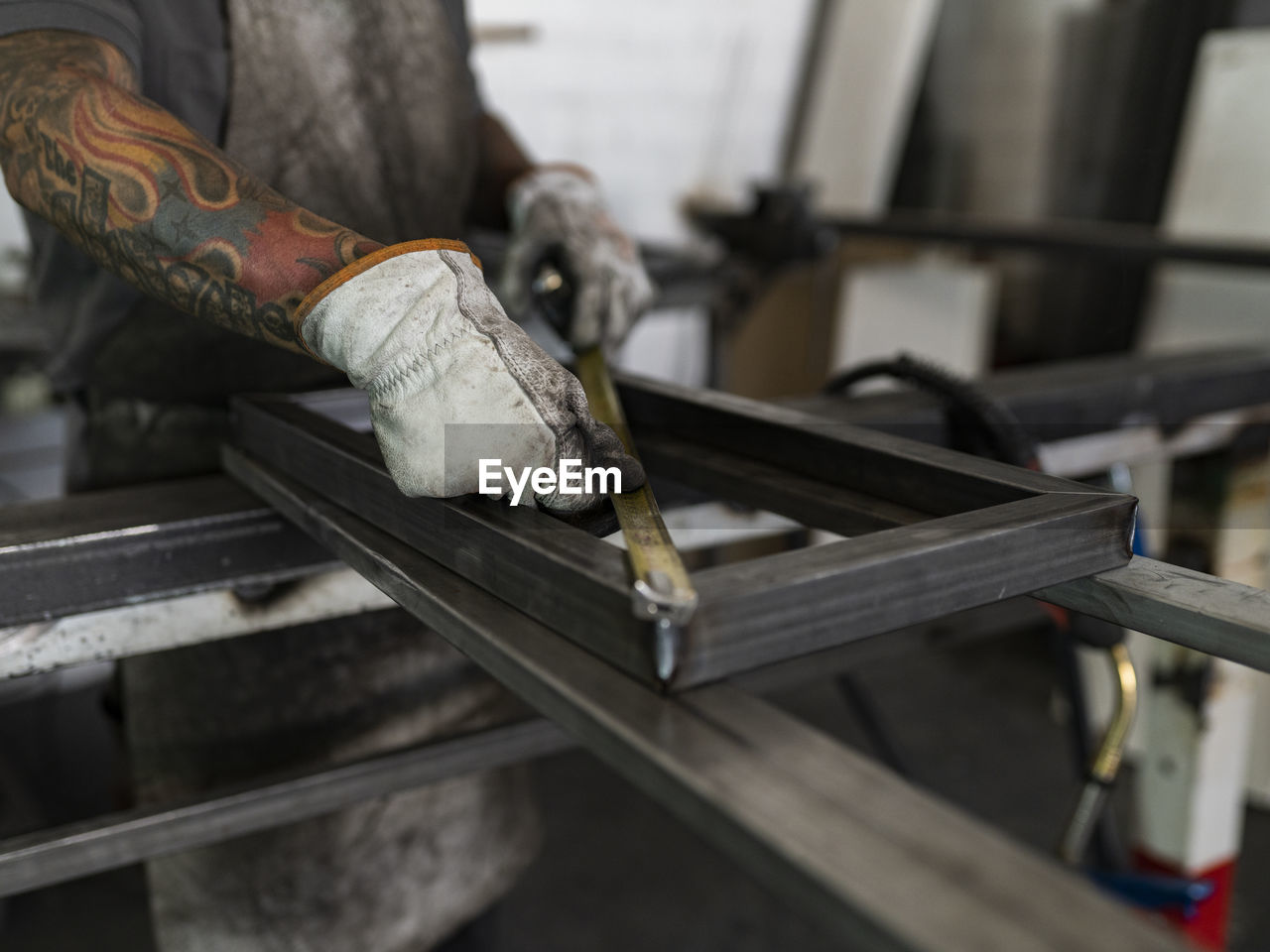 Unrecognizable male welder in dirty gloves and apron measuring frame with yellow metal tape and working at workbench in garage