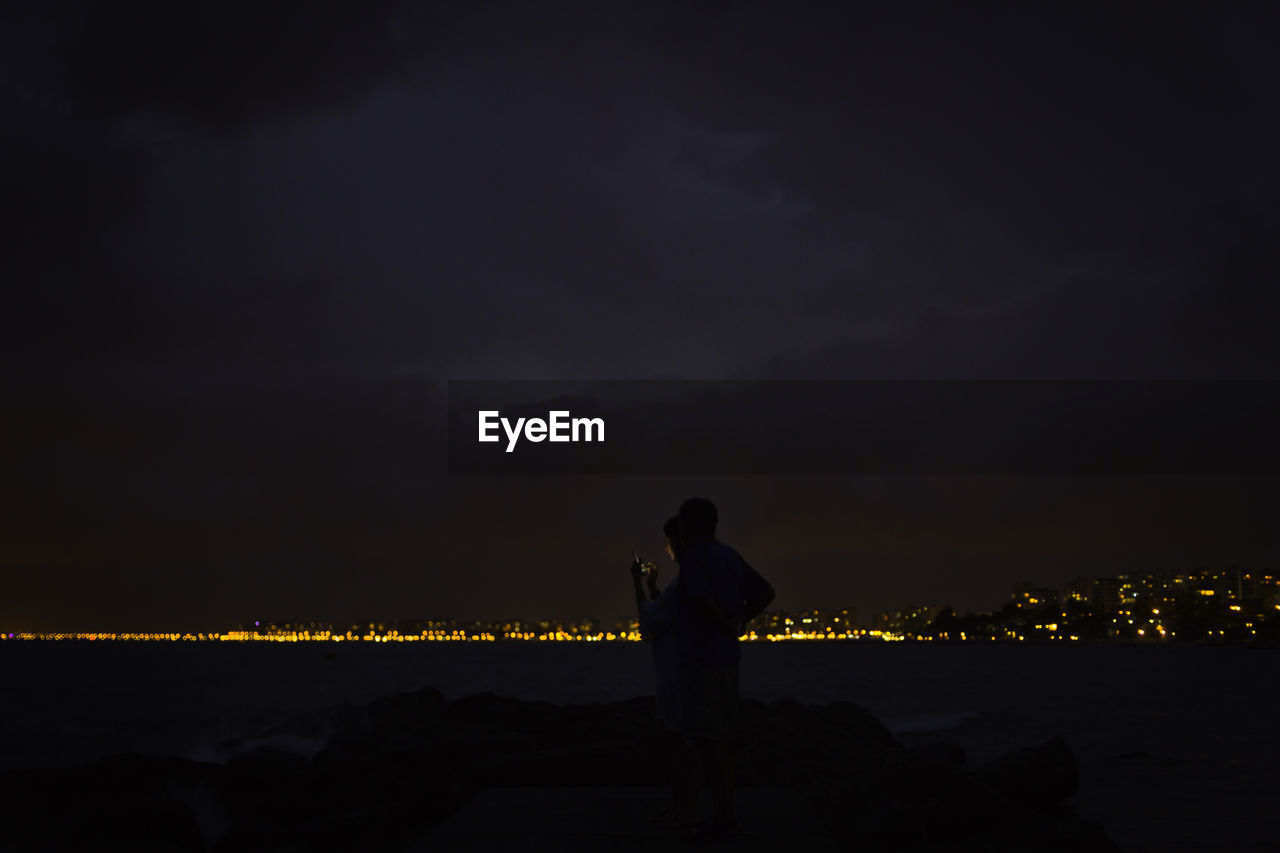 MAN STANDING AT ILLUMINATED SHORE AGAINST SKY DURING SUNSET