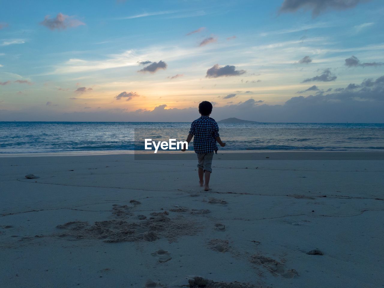 REAR VIEW OF MAN STANDING AT BEACH DURING SUNSET