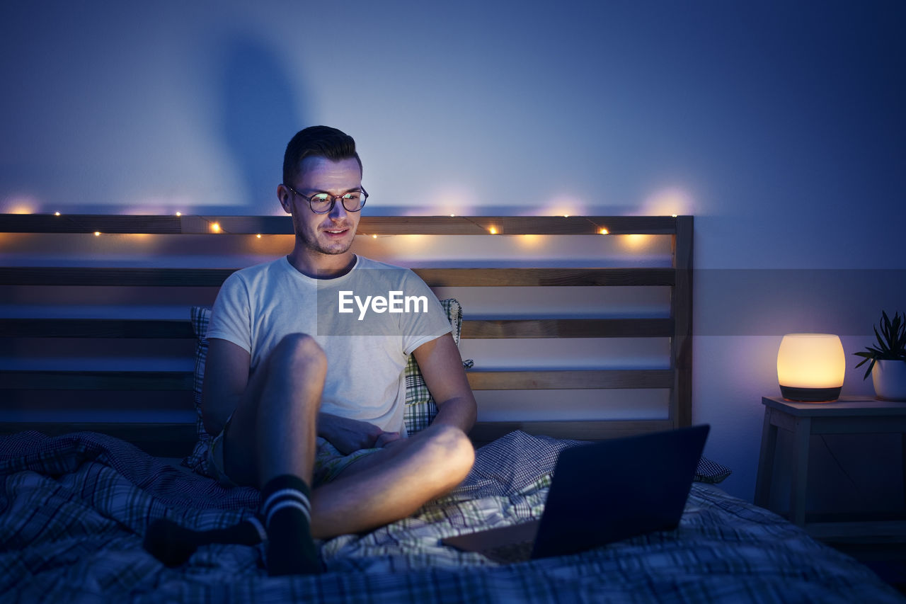 Lonely man with eyeglasses sitting on bed in bedroom and using laptop at night. 