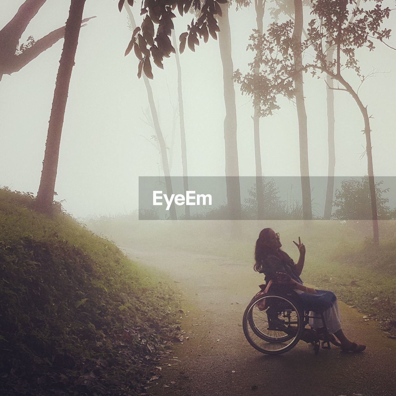 Woman gesturing peace sign while sitting on wheelchair during foggy weather