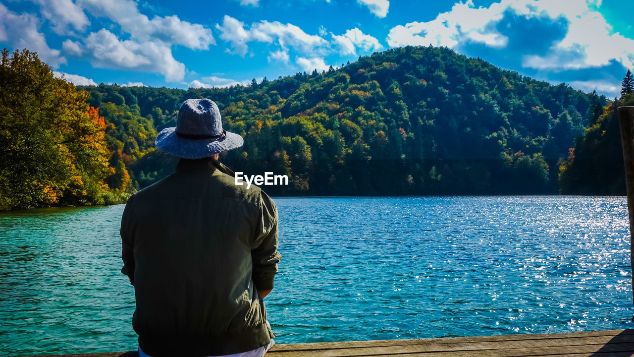 Rear view of man looking at lake while sitting on pier against sky