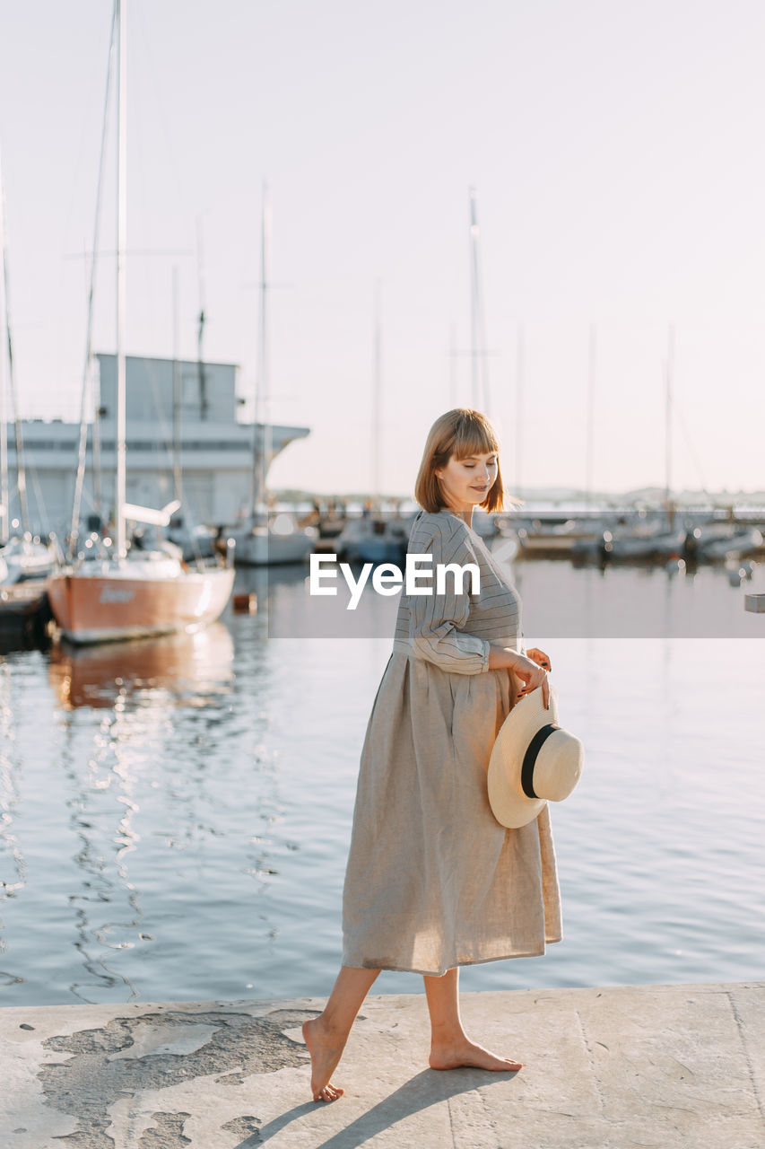 Happy young woman in dress and hat walks on the beach near the boats