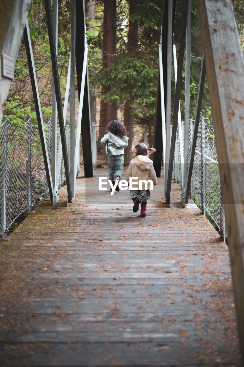 Sisters chase each other over a bridge on a forested path.