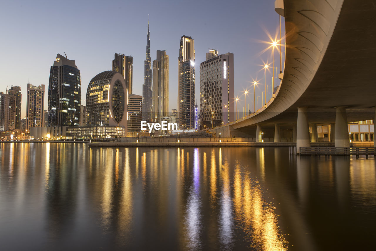 ILLUMINATED BUILDINGS BY RIVER AGAINST SKY AT NIGHT