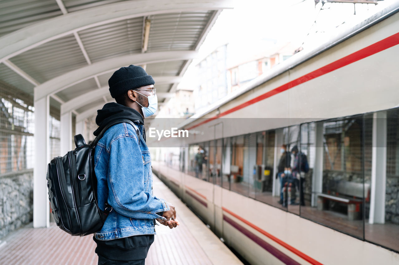 Side view of ethnic male tourist in mask and with backpack standing on platform of railway station during coronavirus epidemic