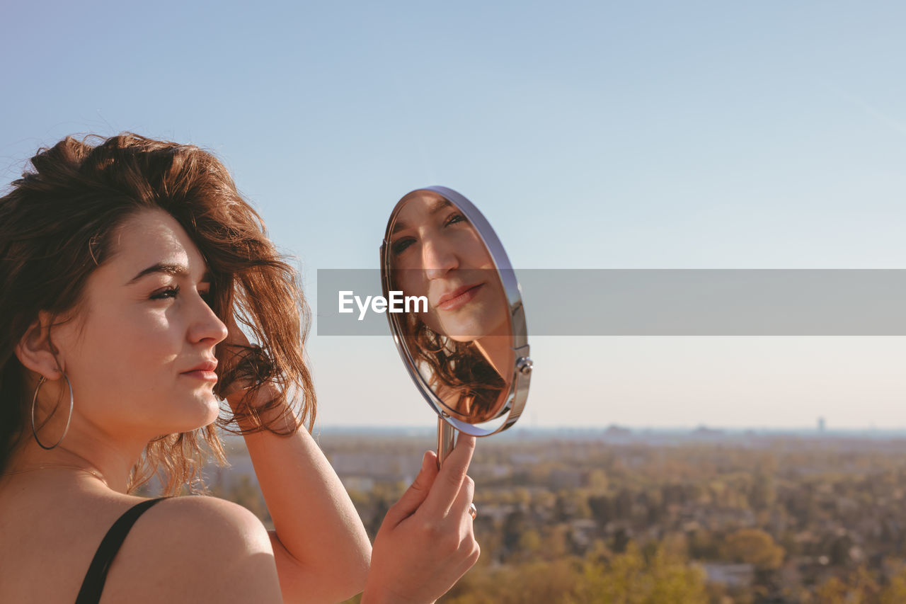 Side view of young woman looking in mirror against clear sky