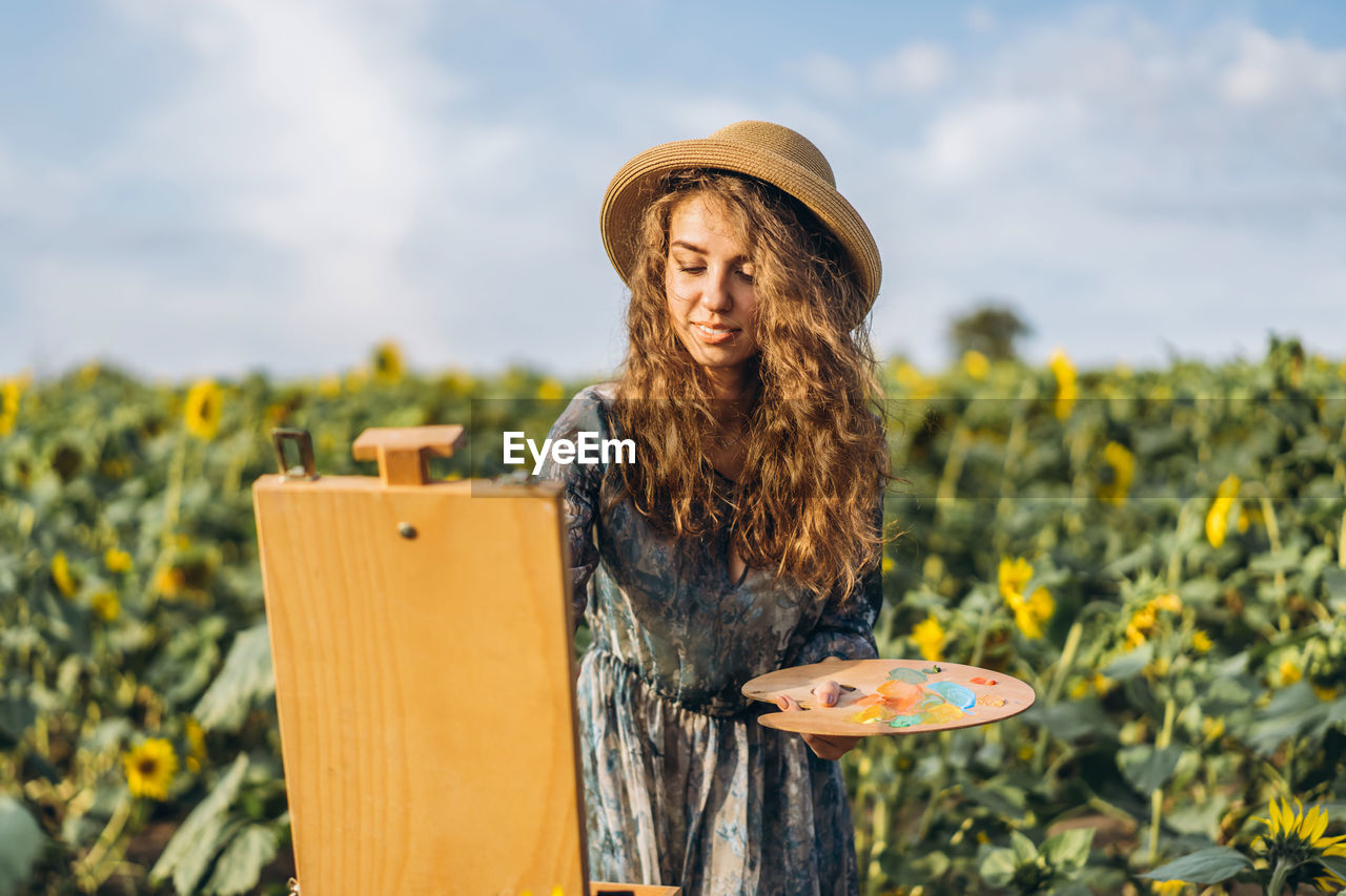 A young woman with curly hair and wearing a hat is painting in nature. 