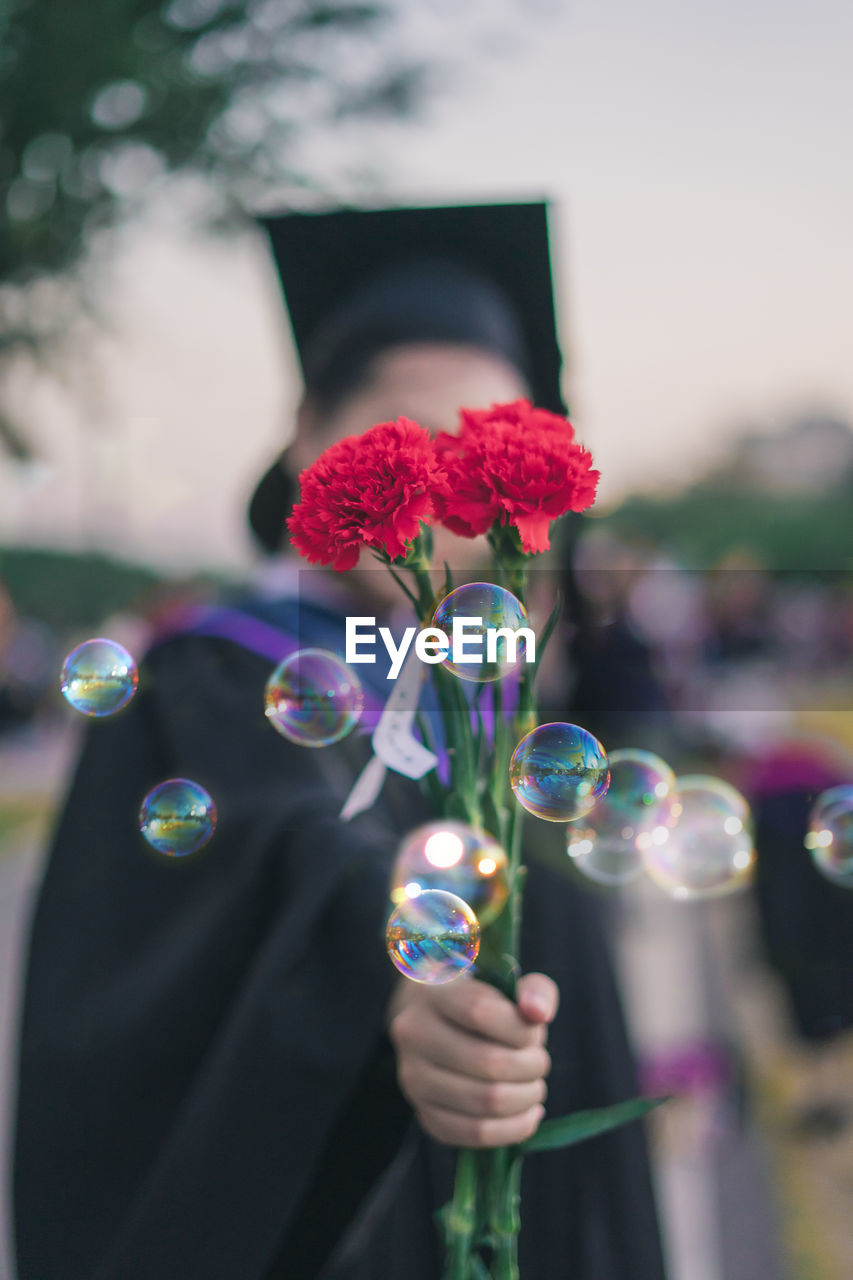 Woman in graduation gown holding red flowers by bubbles