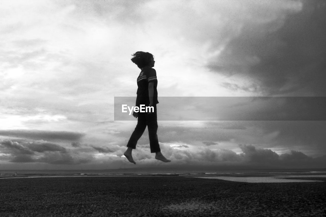 Side view of woman levitating at beach against cloudy sky