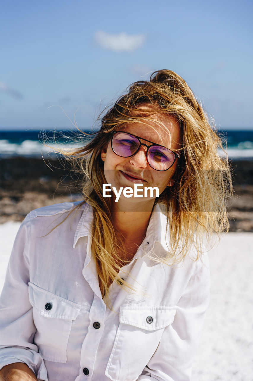 PORTRAIT OF WOMAN WITH SUNGLASSES STANDING AT BEACH