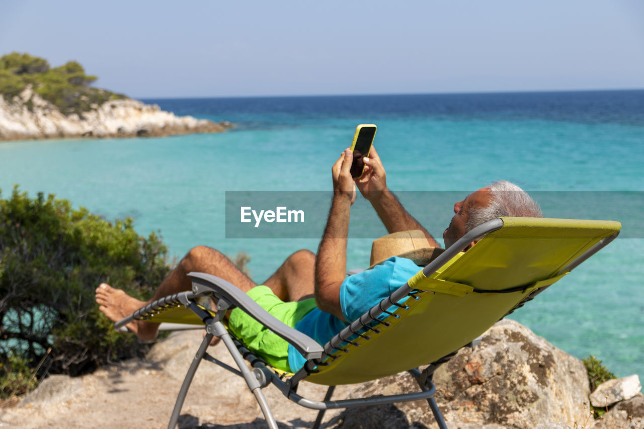 A man on holiday relax in the sun on the deck chair holding his phone chatting with and technology.