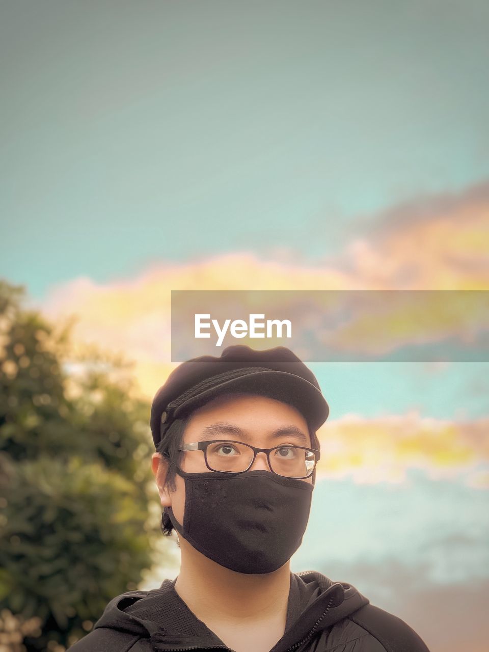 Portrait of young man in cap, eyeglasses and face mask standing against cloudy sunset sky