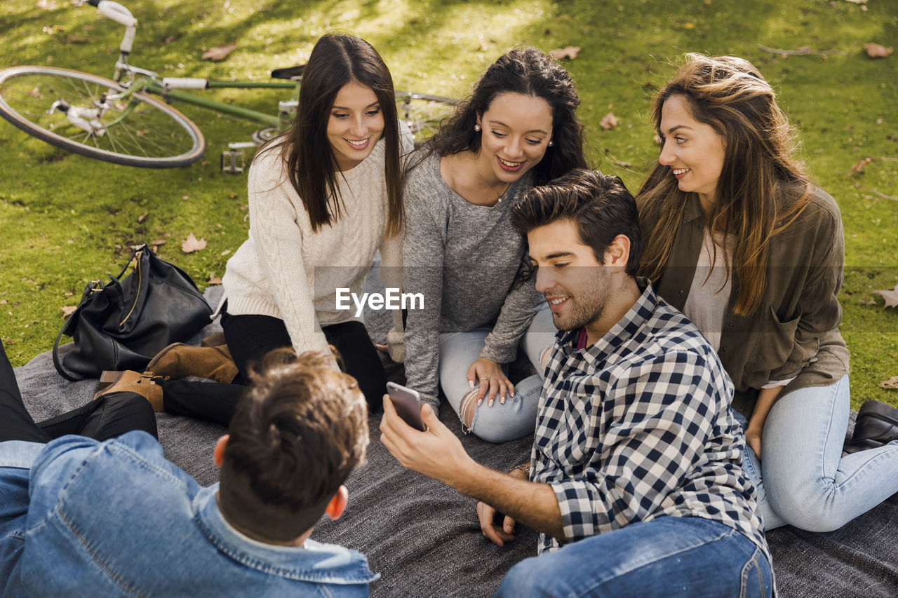 Smiling friends using mobile phone while enjoying picnic at park