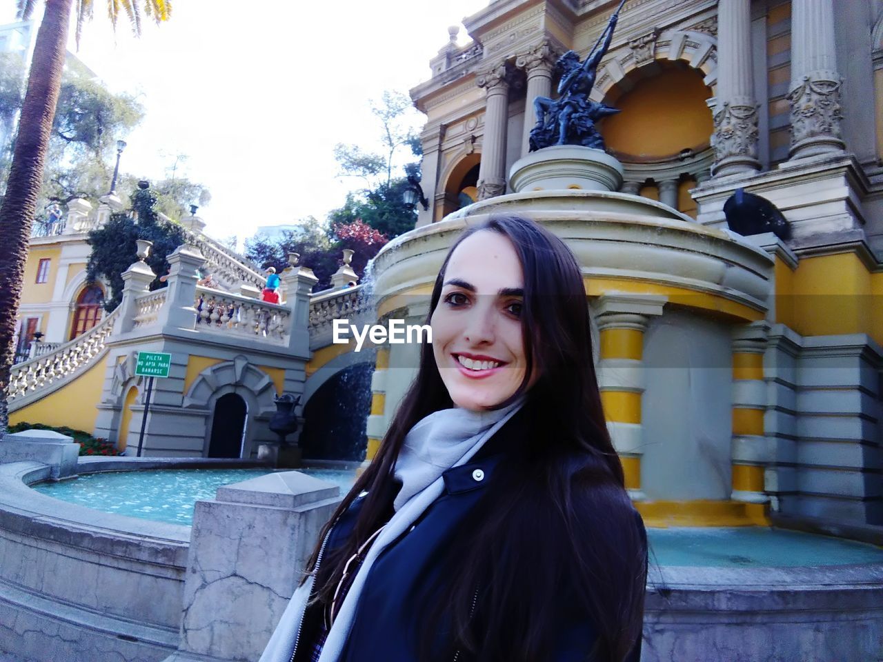 Portrait of smiling young woman against fountain and building