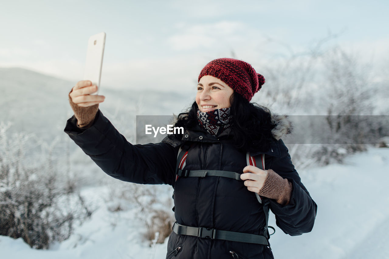 Smiling woman in warm taking selfie while standing on snowcapped mountain