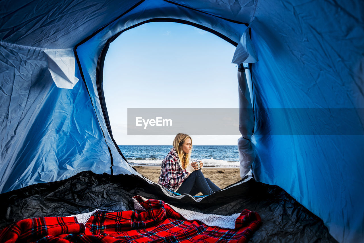 Young woman sitting outside tent by sea