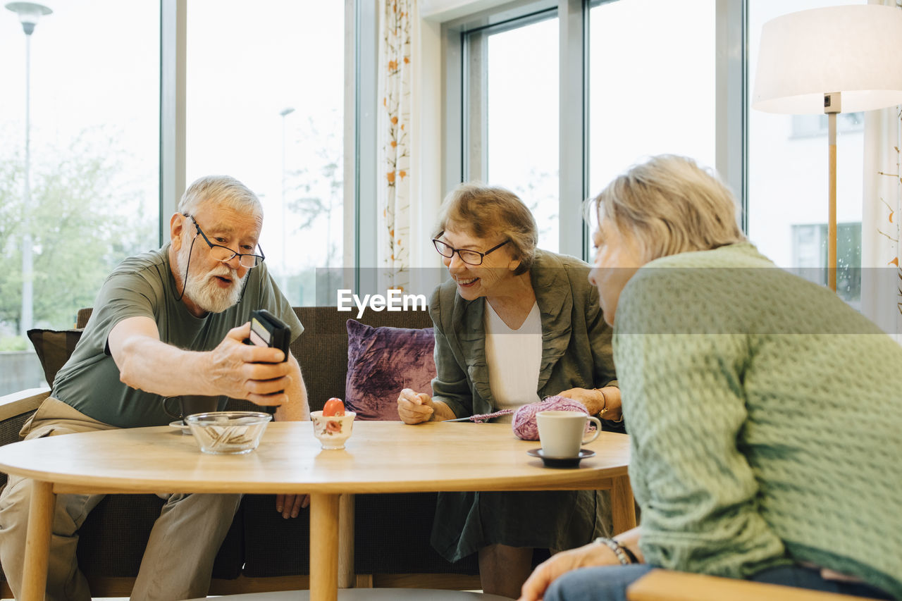 Retired elderly man sharing smart phone with friends sitting at table in nursing home